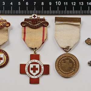 Può includere: Cinque medaglie della Croce Rossa con nastri bianchi e una medaglia d'oro con una croce e uno spillo di sicurezza. Le medaglie sono per la competenza nel lavoro della Croce Rossa e nel pronto soccorso. Il testo sulle medaglie recita "Proficiency in Red Cross Work" e "Proficiency in Red Cross First Aid".