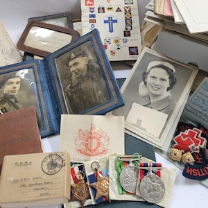 Puede incluir: Una colección de recuerdos militares antiguos, que incluye fotografías enmarcadas, medallas, documentos y insignias. Los artículos están dispuestos sobre una superficie, mostrando una variedad de artefactos históricos y efímeros. La paleta de colores está dominada por tonos sepia, azules y rojos.