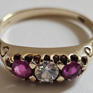 Pode incluir: Um anel de ouro com três pedras preciosas. Duas pedras magenta ladeiam uma pedra clara e facetada. O anel tem um design decorativo nas laterais e um acabamento polido. Uma joia.