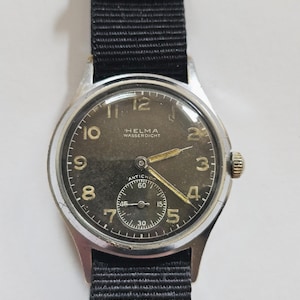 May include: Vintage Helma watch with a black nylon strap. The watch face is dark with gold-colored hands and numerals. The watch has a small seconds dial and the text "HELMA WASSERDICHT" is visible.