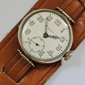 Op de afbeelding: Een zilveren Zenith polshorloge met een witte wijzerplaat en zwarte Romeinse cijfers. Het horloge heeft een bruin leren bandje.