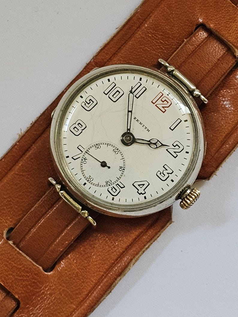 Op de afbeelding: Een zilveren Zenith polshorloge met een witte wijzerplaat en zwarte Romeinse cijfers. Het horloge heeft een bruin leren bandje.