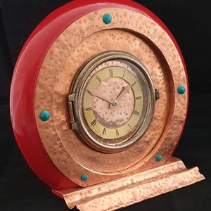 Puede incluir: Un reloj de cobre y rojo con una esfera redonda y detalles turquesa. El reloj está colocado sobre un soporte de cobre.