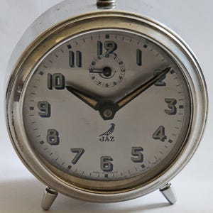 May include: A silver JAZ alarm clock with a circular face and a curved body. The clock face is light-coloured with black numerals and hands. The clock has a small bird logo and the word "JAZ" printed on the face. The clock has three small legs.