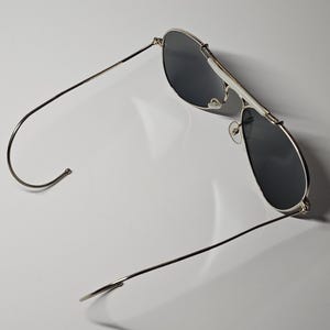 Pu&ograve; includere: Occhiali da sole da aviatore con montatura argentata e lenti scure. Gli occhiali hanno una classica forma a goccia e un doppio ponte. Le aste sono sottili e si estendono fino alle estremit&agrave; curve. Sfondo bianco.