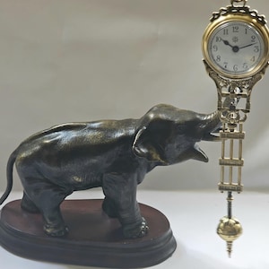 May include: A bronze elephant sculpture with an open mouth, set on a dark wooden base. A decorative clock with a gold frame and a white face is attached to the elephant's trunk. The clock has black Roman numerals.