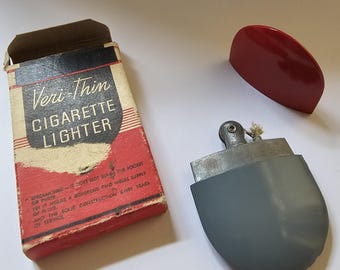 May include: A vintage silver cigarette lighter with a grey plastic case and a red plastic lid. The lighter is in its original box, which is red and white and says "Veri-Thin Cigarette Lighter".