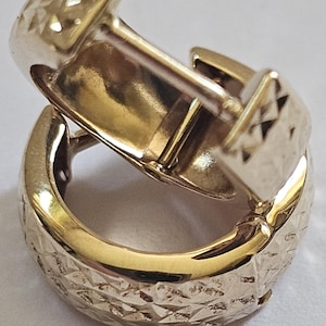 May include: A pair of gold-colored hoop earrings. The earrings have a textured, diamond-cut pattern on the outer surface and a smooth, polished interior. The clasp is visible, and the earrings are set against a white background.