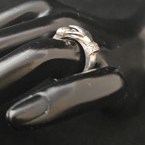 May include: A silver ring featuring a hand design. The ring's hand is sculpted with intricate details, and the band has decorative elements. The ring is shown on a black surface, highlighting its metallic sheen and design.