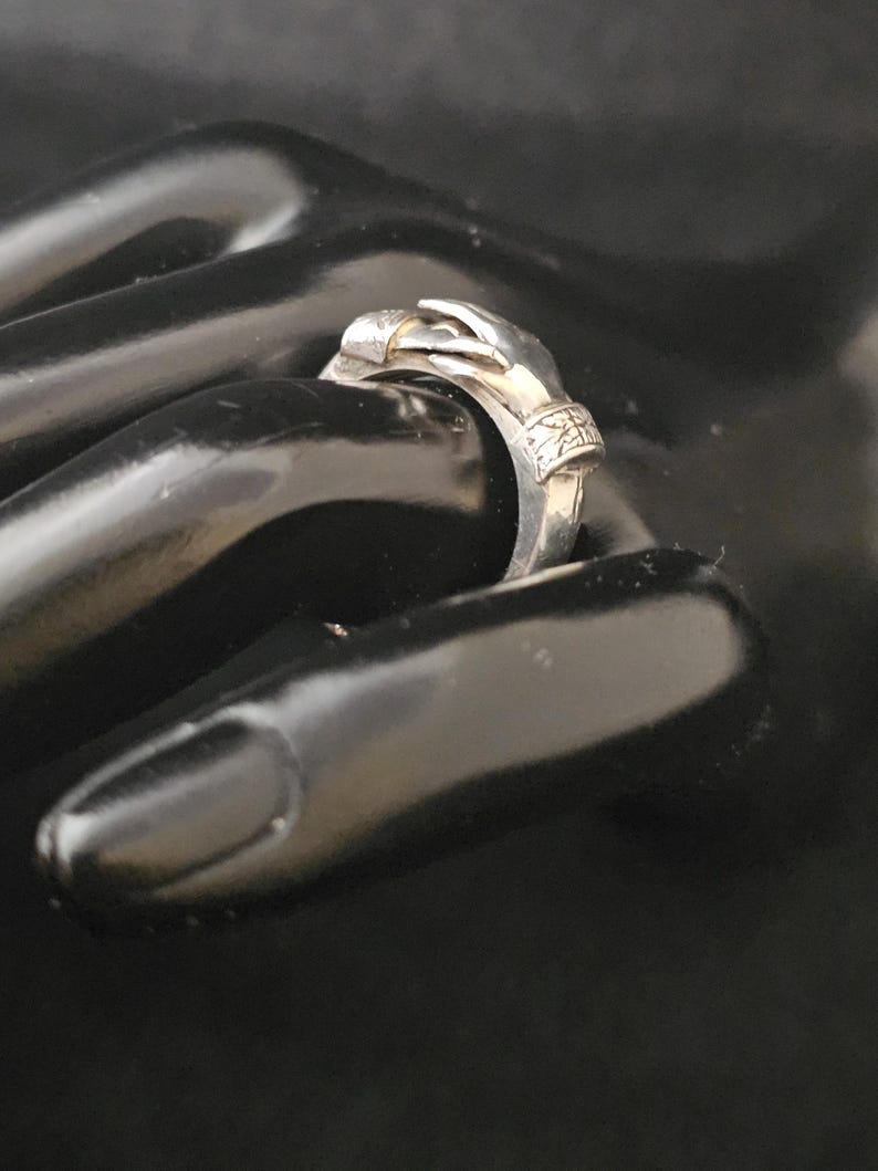 May include: A silver ring featuring a hand design. The ring's hand is sculpted with intricate details, and the band has decorative elements. The ring is shown on a black surface, highlighting its metallic sheen and design.