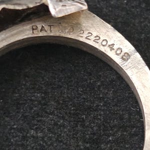 May include: Close-up of a silver metal ring with the text "PAT. NO. 2220408" engraved on the band. The ring has a decorative top with a textured design. The background is a dark, textured surface.
