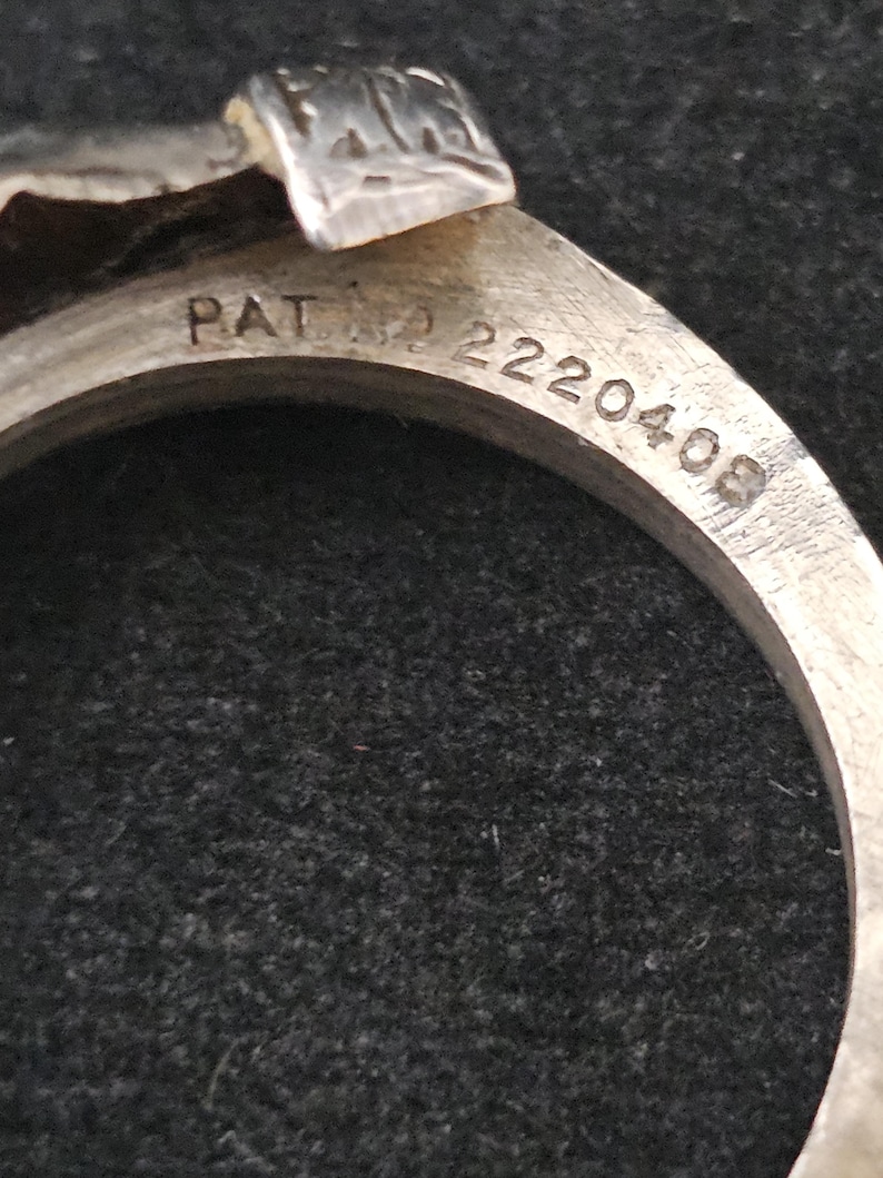 May include: Close-up of a silver metal ring with the text "PAT. NO. 2220408" engraved on the band. The ring has a decorative top with a textured design. The background is a dark, textured surface.