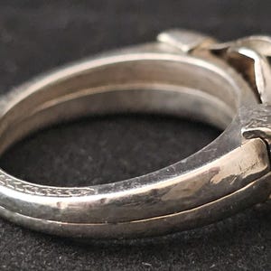 May include: A silver-toned hinged bangle bracelet with a detailed design. The bracelet features a textured clasp and decorative patterns along the edges. The overall design is intricate and appears to be vintage.