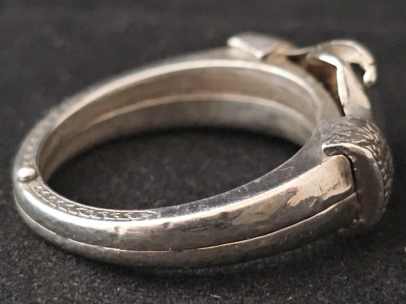May include: A silver-toned hinged bangle bracelet with a detailed design. The bracelet features a textured clasp and decorative patterns along the edges. The overall design is intricate and appears to be vintage.