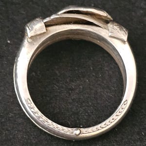 May include: Close-up of a silver ring with a unique design. The ring features a layered, textured top and decorative engravings along the band. The ring is made of a shiny, metallic material.