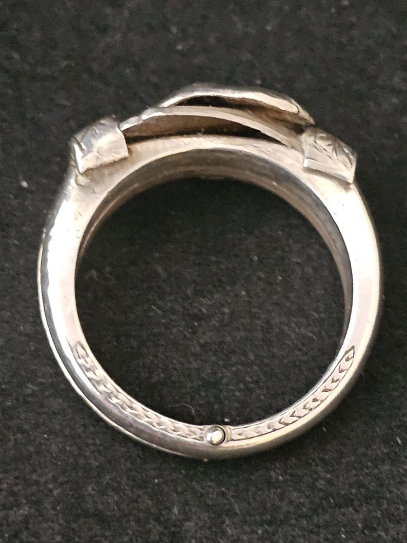 May include: Close-up of a silver ring with a unique design. The ring features a layered, textured top and decorative engravings along the band. The ring is made of a shiny, metallic material.