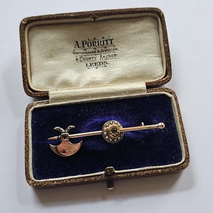 Può includere: Spilla dorata a forma di piccola ascia e design circolare con pietra centrale, montata su una barra d'oro. La spilla è esposta in un cofanetto marrone vintage con il testo "A. PORRITT WATCHMAKER & JEWELLER, 9. COUNTY ARCADE, LEEDS."