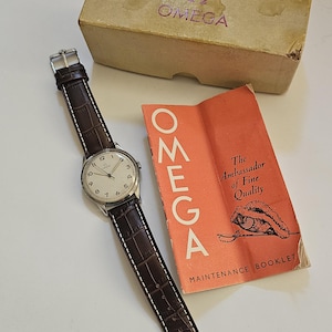 Puede incluir: Un reloj de pulsera Omega vintage con una correa de cuero marrón y una caja plateada. El reloj tiene una esfera blanca con números y agujas negras. La imagen también muestra la caja original de Omega y un folleto de mantenimiento.