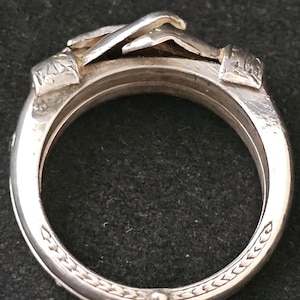 May include: A silver ring with a unique design. The ring features a detailed, raised design of hands clasped together at the top. The band has an intricate pattern around the edges. The ring is made of a shiny, metallic material.