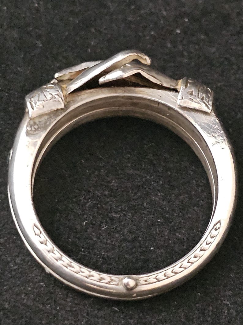 May include: A silver ring with a unique design. The ring features a detailed, raised design of hands clasped together at the top. The band has an intricate pattern around the edges. The ring is made of a shiny, metallic material.