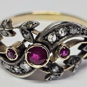 Op de afbeelding: Een antieke ring met een gouden band en een sierlijk zilveren filigraan ontwerp. De ring heeft drie grote, ronde, dieprode robijnen en verschillende kleine, heldere diamanten. Het ontwerp bevat blad- en wijnrankmotieven.