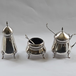 Op de afbeelding: Een set van drie zilveren zout- en peperstrooiers met sierlijke ontwerpen en een klein lepeltje. De strooiers hebben een koepelvormig deksel en staan op drie poten.