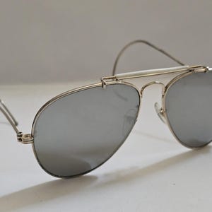 Pu&ograve; includere: Occhiali da sole da aviatore argentati con lenti a specchio. Gli occhiali hanno una montatura argentata e un ponte bianco. Le lenti sono di colore argento riflettente. Le aste sono sottili e argentate.