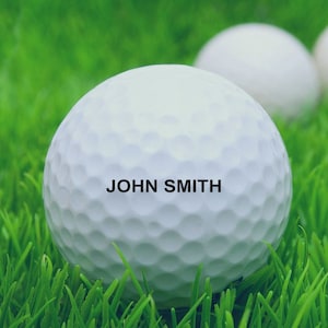 May include: A white golf ball with the name "JOHN SMITH" printed on it, sitting on green grass.