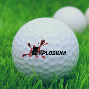 May include: A white golf ball with a red and black logo that says "EXPLOSIUM" on it. The golf ball is sitting on green grass.