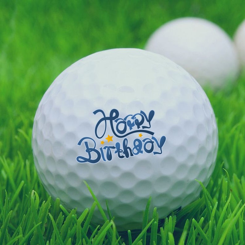 May include: A white golf ball with the words "Happy Birthday" written in blue and yellow on it. The golf ball is sitting on green grass.