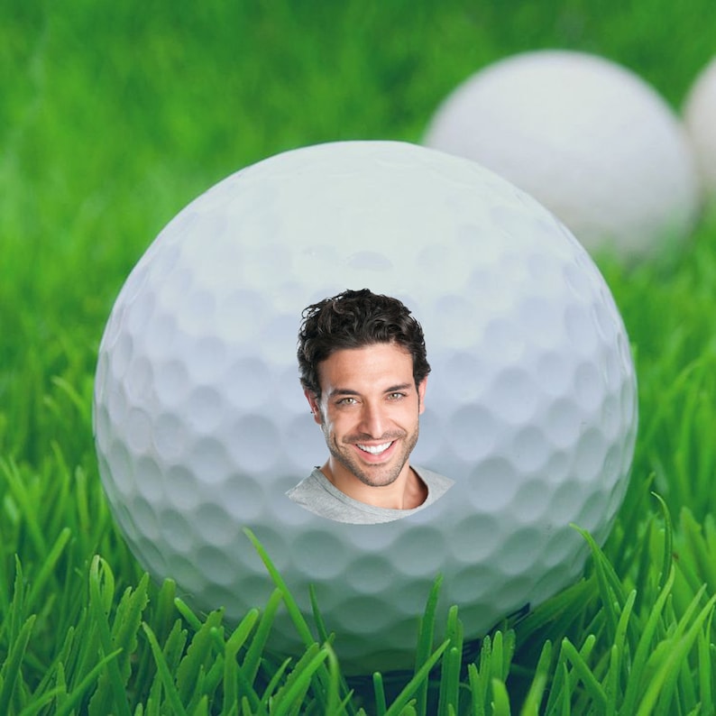 May include: A white golf ball with a man's face peeking through a hole in the center. The ball is sitting on green grass.