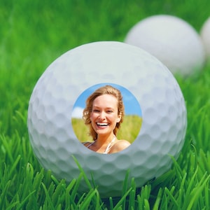 May include: A white golf ball with a smiling woman's face in the center of the ball. The ball is sitting on green grass.