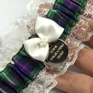 May include: White lace garter with a green, blue, and red plaid ribbon and a white satin bow. A silver charm with the text "YOUR ENGRAVING HERE" is attached to the bow.