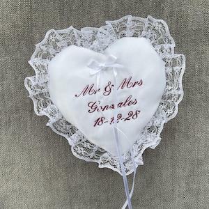 May include: A white heart-shaped ring pillow with a delicate lace trim. The pillow features embroidered text in burgundy that reads "Mr & Mrs Gonzales 18-12-28". A small white bow adorns the top, and white ribbons hang from the bottom.