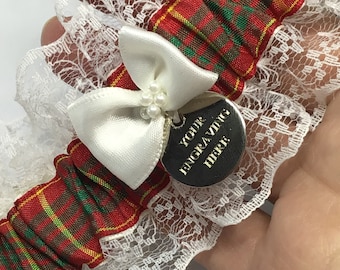 May include: A red, green, and yellow plaid garter with white lace trim and a white satin bow. A silver charm with the text "YOUR ENGRAVING HERE" is attached to the bow.