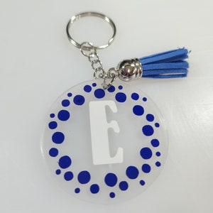 Pode incluir: Um chaveiro de acr&iacute;lico transparente com um padr&atilde;o de bolinhas azuis e uma letra "E" branca no centro. O chaveiro tem um anel de metal prateado e um tassel azul.