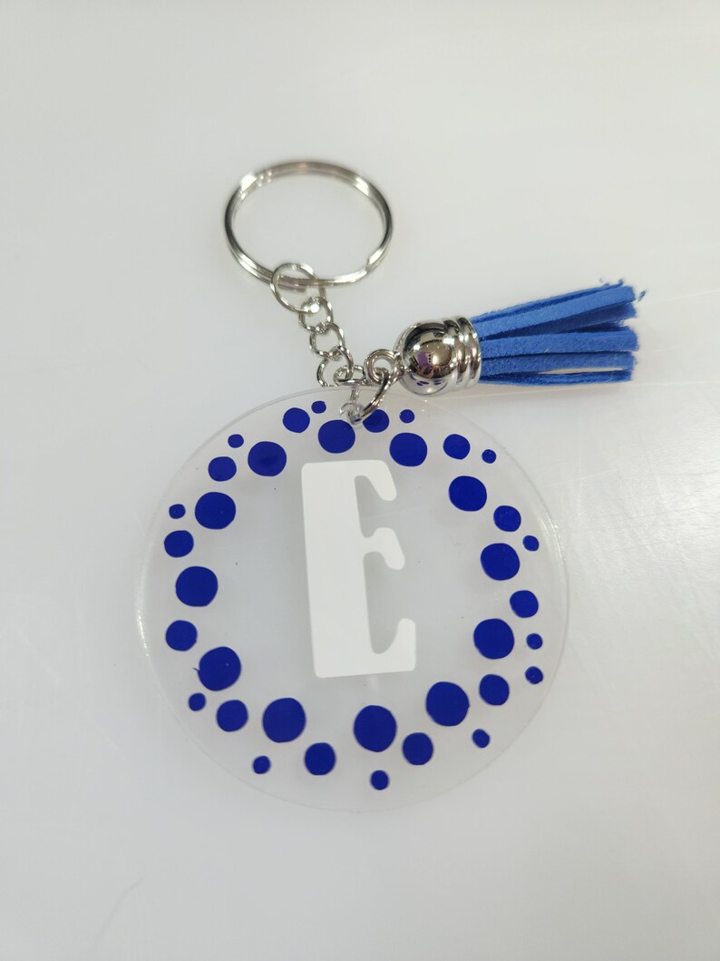 Pode incluir: Um chaveiro de acr&iacute;lico transparente com um padr&atilde;o de bolinhas azuis e uma letra "E" branca no centro. O chaveiro tem um anel de metal prateado e um tassel azul.