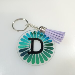 Pode incluir: Um chaveiro iridescente azul-turquesa com uma letra D preta no centro e um tassel lavanda.