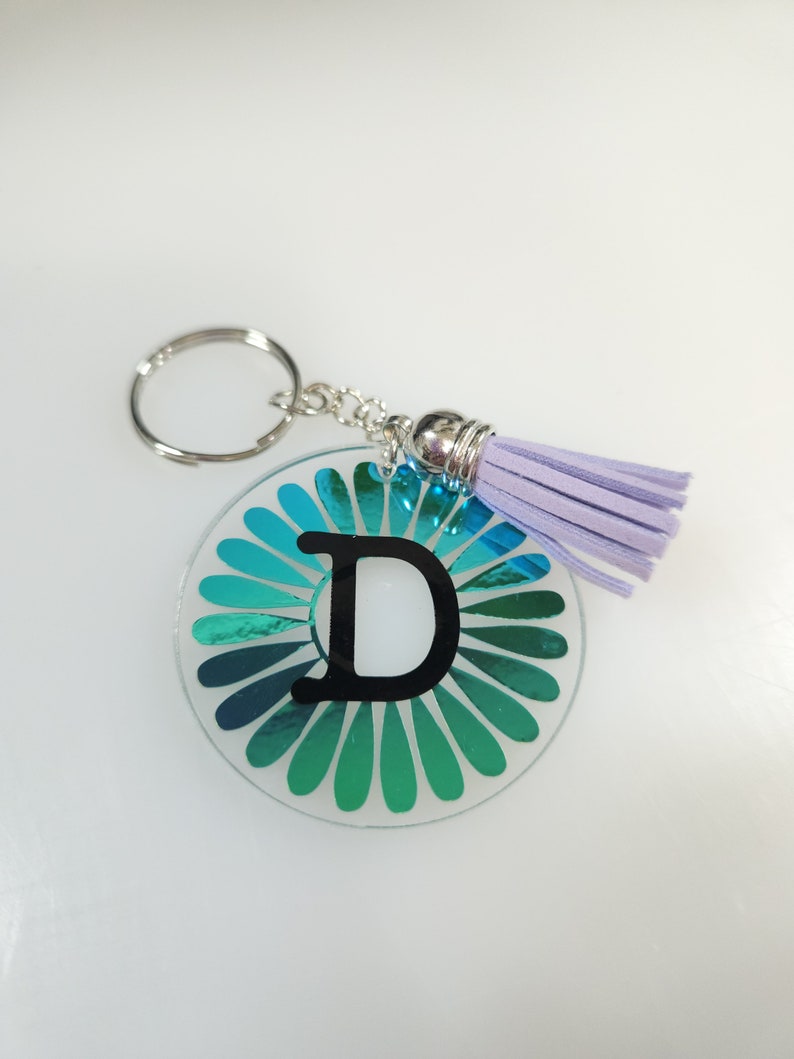 Pode incluir: Um chaveiro iridescente azul-turquesa com uma letra D preta no centro e um tassel lavanda.