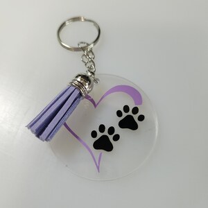 Pode incluir: Um chaveiro de acr&iacute;lico transparente com um cora&ccedil;&atilde;o roxo e duas pegadas pretas. O chaveiro tem um tassel roxo.