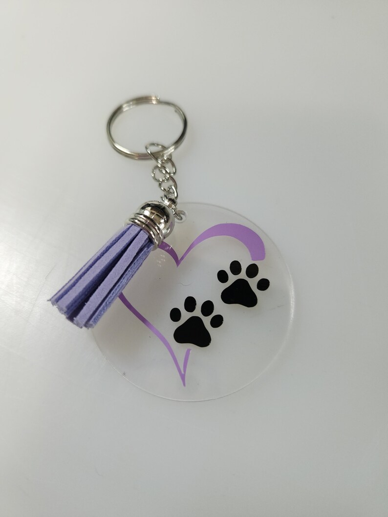Pode incluir: Um chaveiro de acr&iacute;lico transparente com um cora&ccedil;&atilde;o roxo e duas pegadas pretas. O chaveiro tem um tassel roxo.