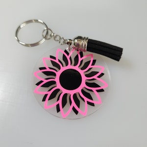 Pode incluir: Um chaveiro de girassol rosa e preto com um tassel preto. O girassol &eacute; contornado em rosa e tem um centro preto.