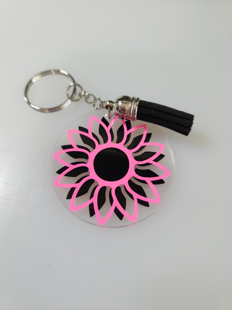 Pode incluir: Um chaveiro de girassol rosa e preto com um tassel preto. O girassol &eacute; contornado em rosa e tem um centro preto.