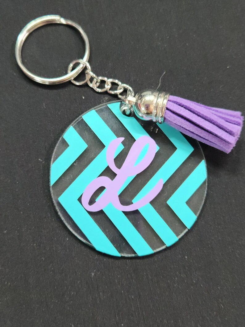 Pode incluir: Um chaveiro de acr&iacute;lico com padr&atilde;o turquesa e preto com uma letra "L" roxa e um tassel roxo.