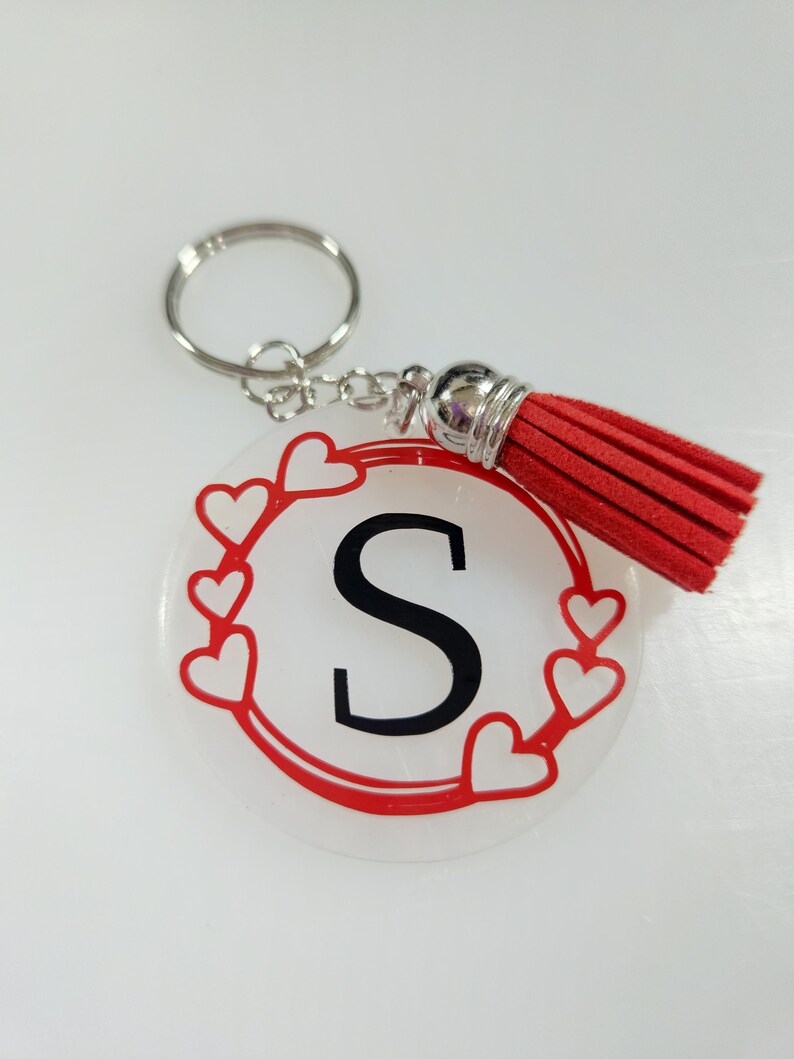 Pode incluir: Um chaveiro de acr&iacute;lico transparente com uma borda vermelha e cora&ccedil;&otilde;es brancos. A letra "S" est&aacute; em preto dentro do c&iacute;rculo. Um tassel vermelho pendura do chaveiro.