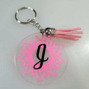 Pode incluir: Um chaveiro de acr&iacute;lico transparente e rosa com um tassel rosa. O chaveiro apresenta um design de mandala rosa e a letra "G" em preto.