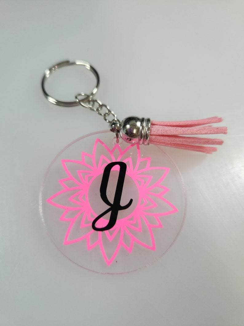 Pode incluir: Um chaveiro de acr&iacute;lico transparente e rosa com um tassel rosa. O chaveiro apresenta um design de mandala rosa e a letra "G" em preto.