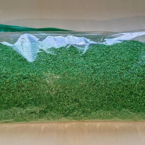 May include: A clear plastic bag filled with bright green, finely cut material. The bag has a green zipper closure and is partially transparent, allowing the contents to be visible. The material appears to be a dried herb or spice.