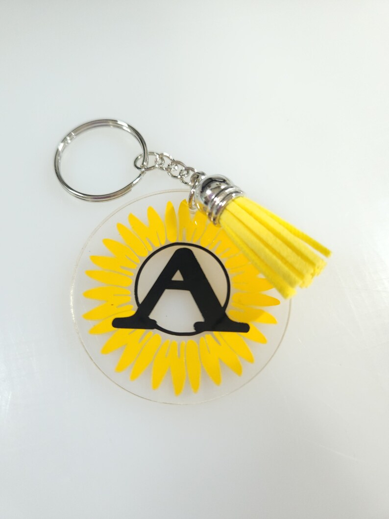 Pode incluir: Um chaveiro de girassol amarelo com uma letra A preta no centro. O chaveiro tem um anel prateado e um tassel amarelo.