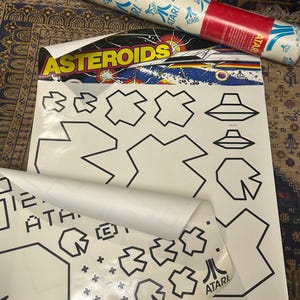May include: A white sheet with black outlines of geometric shapes and the word "ASTEROIDS" in yellow and red. The Atari logo is visible. Rolled-up posters with the Atari logo are in the background. The sheet has a retro video game theme.